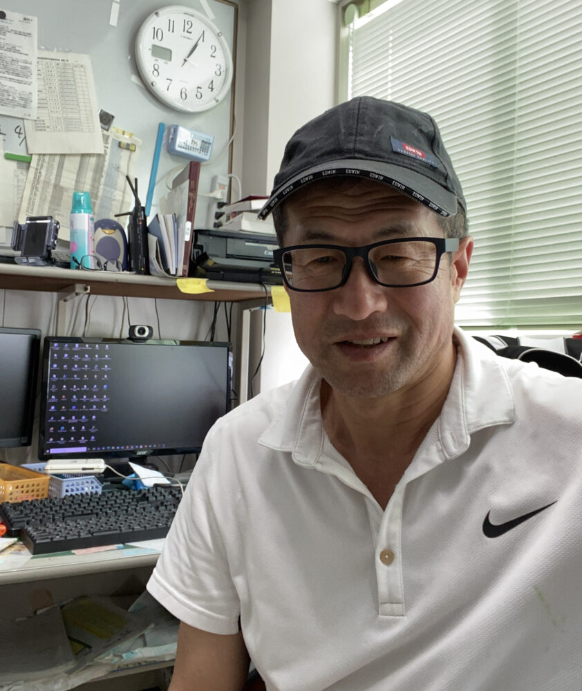 研究室で、ポロシャツとキャップをかぶった小野先生